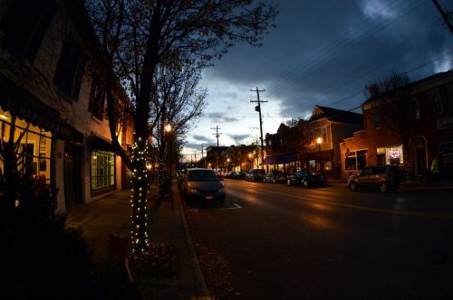 A quiet street at dusk, lined with shops and trees, illuminated by streetlights and a cloudy sky.