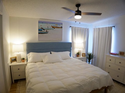 A cozy bedroom with a large bed, blue headboard, two lamps, and a painting of boats on the wall. Bright and inviting decor.