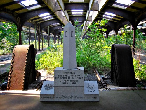 A stone monument honoring Central Railroad employees, surrounded by overgrown vegetation in an abandoned train station.