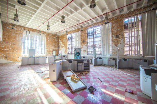 An abandoned office space with scattered furniture, large windows, and sunlight streaming through.