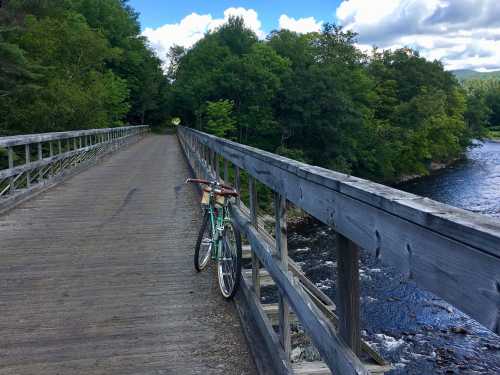 A bicycle rests on a wooden bridge overlooking a river, surrounded by lush green trees and a blue sky.