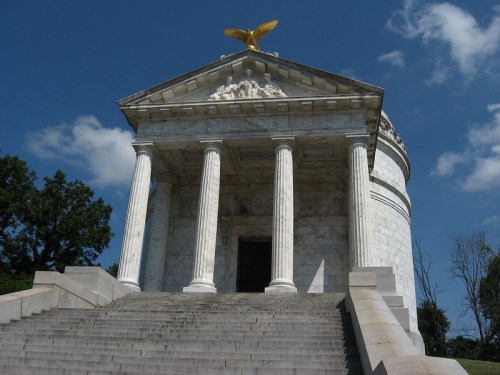 A marble temple with columns and a golden eagle atop, set against a blue sky and surrounded by trees.