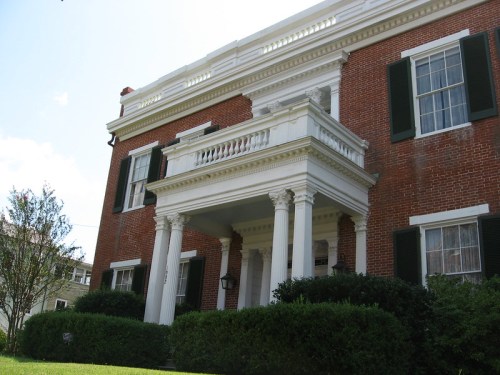 A large brick house with a white columned porch and green shutters, surrounded by manicured bushes and grass.