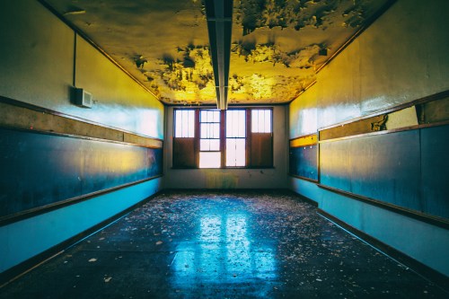 Abandoned classroom with peeling paint, dusty floor, and large windows letting in soft light.