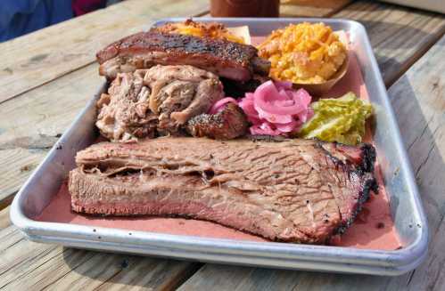 A metal tray filled with smoked meats, including brisket, pulled pork, pickled onions, and sides.