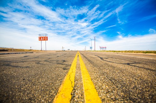 A long, empty road stretches ahead, with a "CAFE" sign visible against a bright blue sky and scattered clouds.