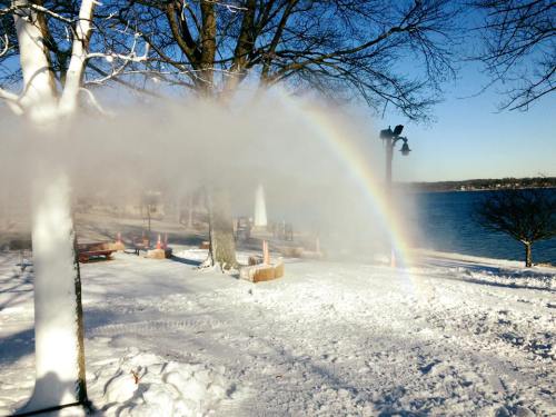 A snowy landscape with trees, a rainbow forming in mist, and a lake in the background under a clear blue sky.