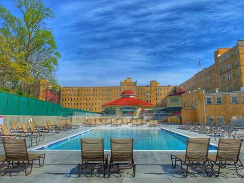A sunny outdoor pool area with lounge chairs, surrounded by a large building and trees under a blue sky.