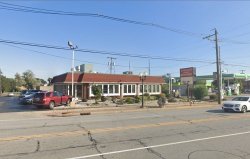 A family restaurant with a red roof, surrounded by greenery, located near a gas station on a busy street.