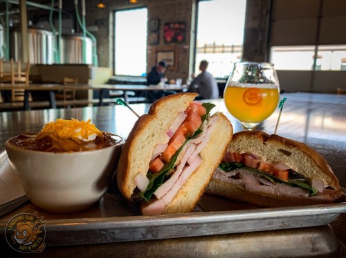 A plate with two sandwiches filled with turkey, lettuce, and tomato, alongside a bowl of chili and a glass of beer.