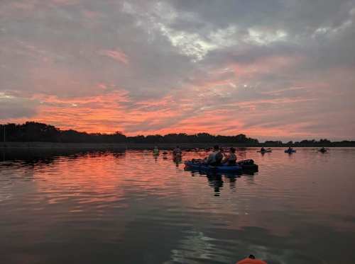 Kayakers on a calm lake at sunset, with vibrant orange and pink hues reflecting on the water.