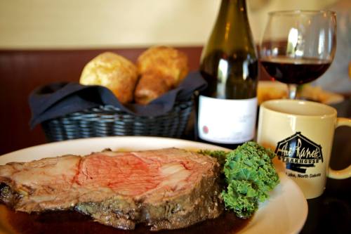 A plate of medium-rare prime rib with kale, accompanied by a glass of red wine and a basket of rolls in the background.