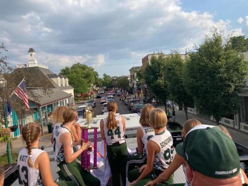 A group of girls in sports jerseys sit on a float, overlooking a parade down a tree-lined street.