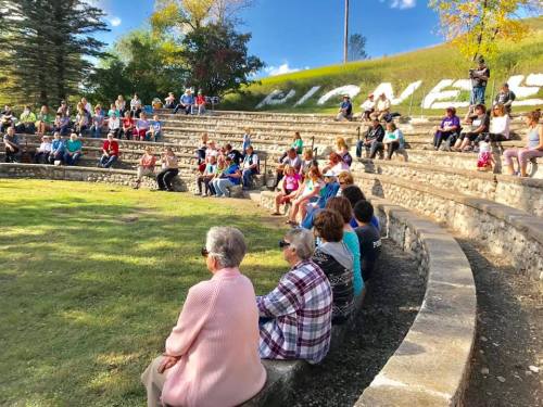 A diverse crowd sits on stone steps in an outdoor amphitheater, enjoying a sunny day surrounded by greenery.