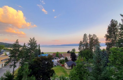 A serene lakeside view at sunset, with trees, houses, and soft clouds reflecting warm colors in the sky.