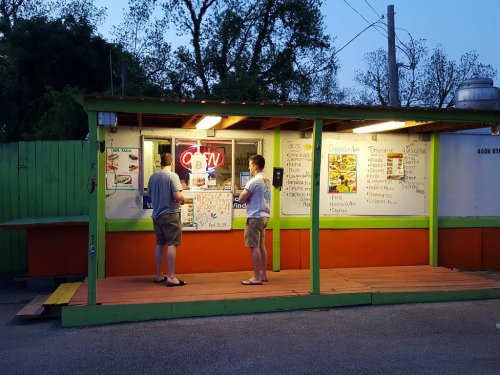 Two people stand at a food stand with an "OPEN" sign, reviewing the menu on the wall in the evening light.