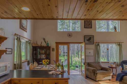 Cozy interior of a cabin with wooden beams, a dining area, and large windows overlooking a forested landscape.