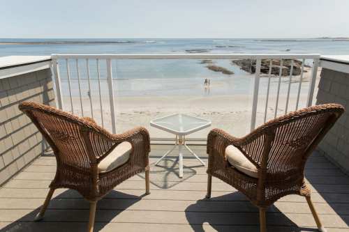 Two wicker chairs and a small table on a balcony overlooking a calm beach and ocean.