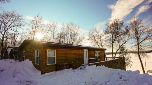 A wooden cabin surrounded by snow, with trees and a lake in the background under a clear blue sky.