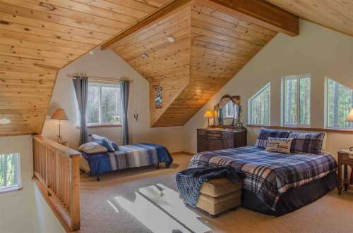 Cozy attic bedroom with wooden beams, two beds, a dresser, and large windows letting in natural light.