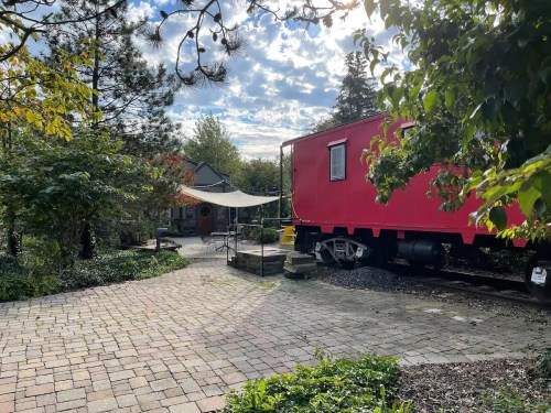 A red train car sits beside a paved path, surrounded by greenery and trees under a partly cloudy sky.