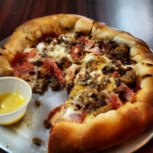 A partially eaten pizza with a stuffed crust, topped with meats and cheese, alongside a small cup of dipping sauce.