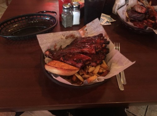A plate of barbecue ribs with sauce, fries, and a bun on a table, with condiments and a drink in the background.