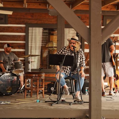 A musician sings on stage with a band, featuring drums and guitar, in a rustic wooden setting.