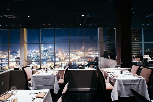 Elegant restaurant interior with tables set for dining, featuring a panoramic city skyline view at night.
