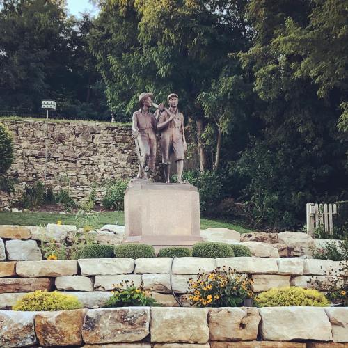 A bronze statue of two figures standing together on a stone pedestal, surrounded by greenery and flowers.