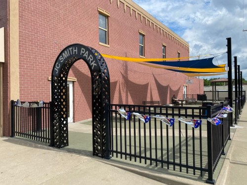Entrance to JC Smith Park featuring a decorative arch and colorful shade sails above a fenced area.
