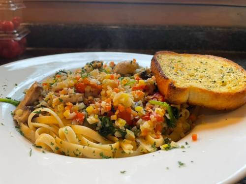 A plate of pasta with mixed vegetables and a slice of garlic bread on the side.