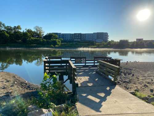 A serene riverside view with a wooden dock, lush greenery, and a building in the background under a clear blue sky.