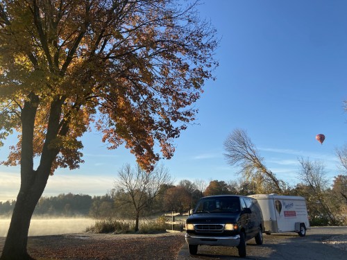 A black van parked near a lake with autumn trees, a trailer, and a hot air balloon in the clear blue sky.