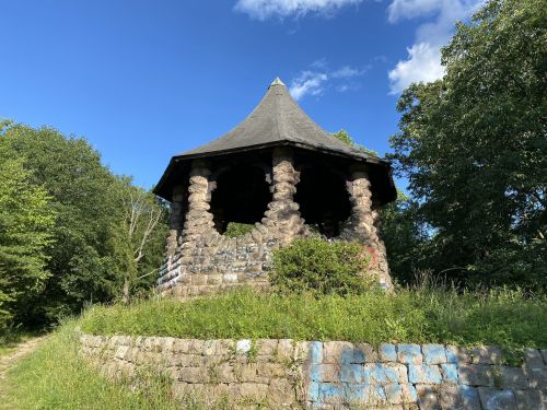 A stone gazebo with a conical roof, surrounded by greenery and blue sky, sits on a grassy hill.