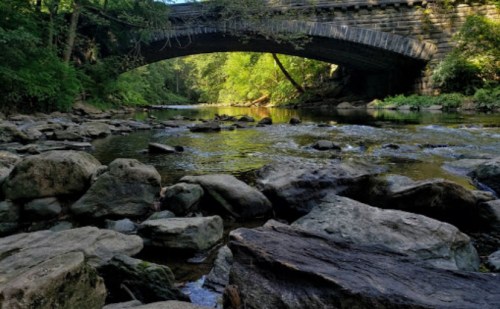 A stone bridge arches over a calm river, surrounded by lush greenery and rocky riverbanks.