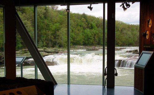 View of a river with a waterfall, framed by large glass windows and surrounded by lush green trees.