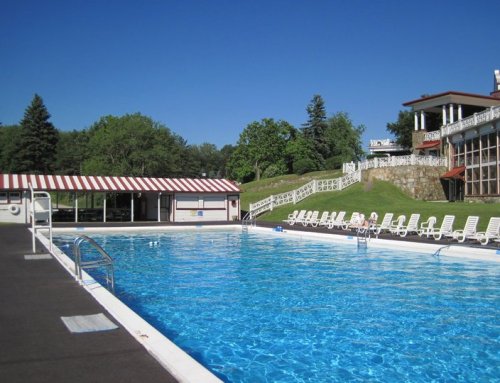 A clear blue swimming pool surrounded by lounge chairs and a building with a striped awning, set in a green landscape.