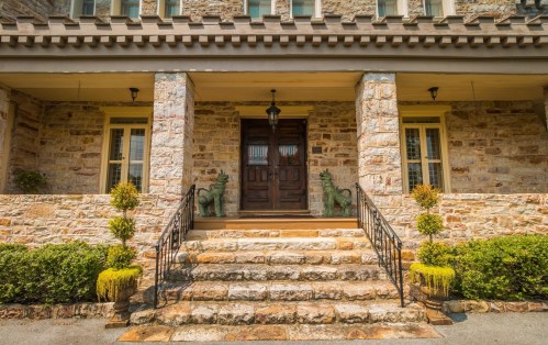 Front entrance of a stone house with steps, two green statues, and potted plants on either side.