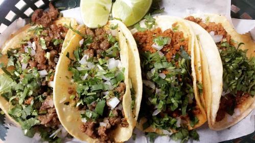 Four tacos filled with seasoned meat, topped with chopped onions and cilantro, served with lime wedges.
