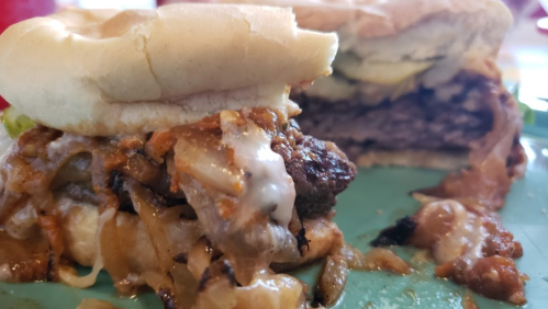 Close-up of a burger with melted cheese, grilled onions, and pickles, served on a soft bun.