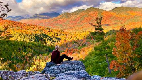 A person sits on a rock, overlooking a vibrant autumn landscape with colorful trees and mountains in the background.