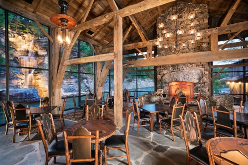 Cozy dining area with wooden beams, stone fireplace, and large windows showcasing a waterfall view.