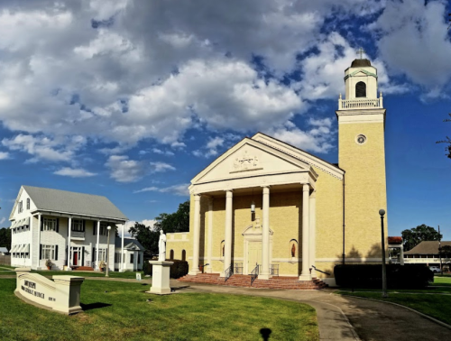 A yellow church with columns and a clock tower, alongside a white building, under a partly cloudy blue sky.
