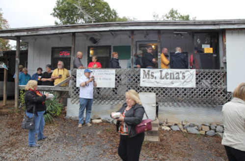 A crowd gathers outside Ms. Lena's restaurant, with people chatting and enjoying food on a cloudy day.