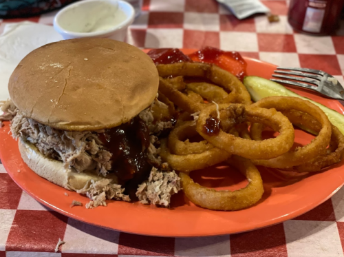 A plate with a pulled pork sandwich, onion rings, pickles, and a side of red sauce on a checkered tablecloth.