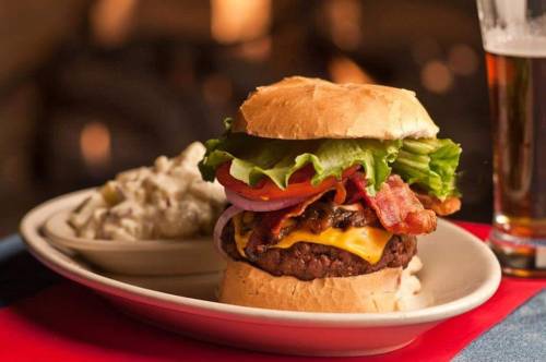 A juicy cheeseburger with bacon, lettuce, tomato, and onion, served with a side of potato salad and a glass of beer.