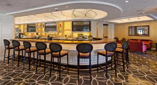A stylish bar with high stools, illuminated shelves, and a cozy seating area in a modern lounge.