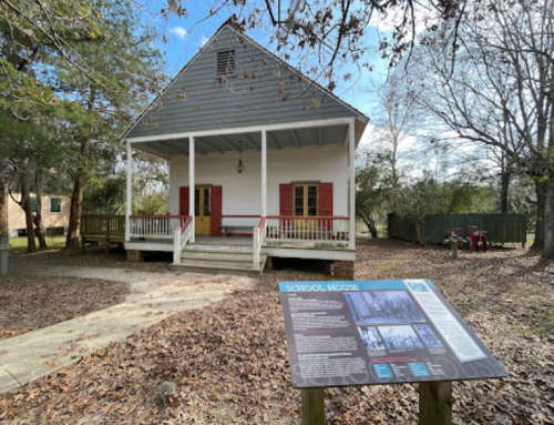A historic schoolhouse with a porch, red shutters, and an informational sign, surrounded by trees and fallen leaves.