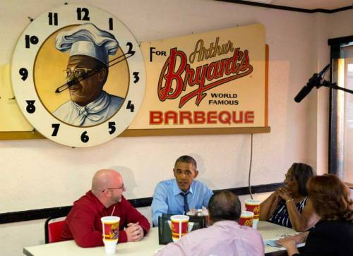 President Obama dining with a group at Arthur Bryant's Barbeque, with a large clock and chef mural in the background.